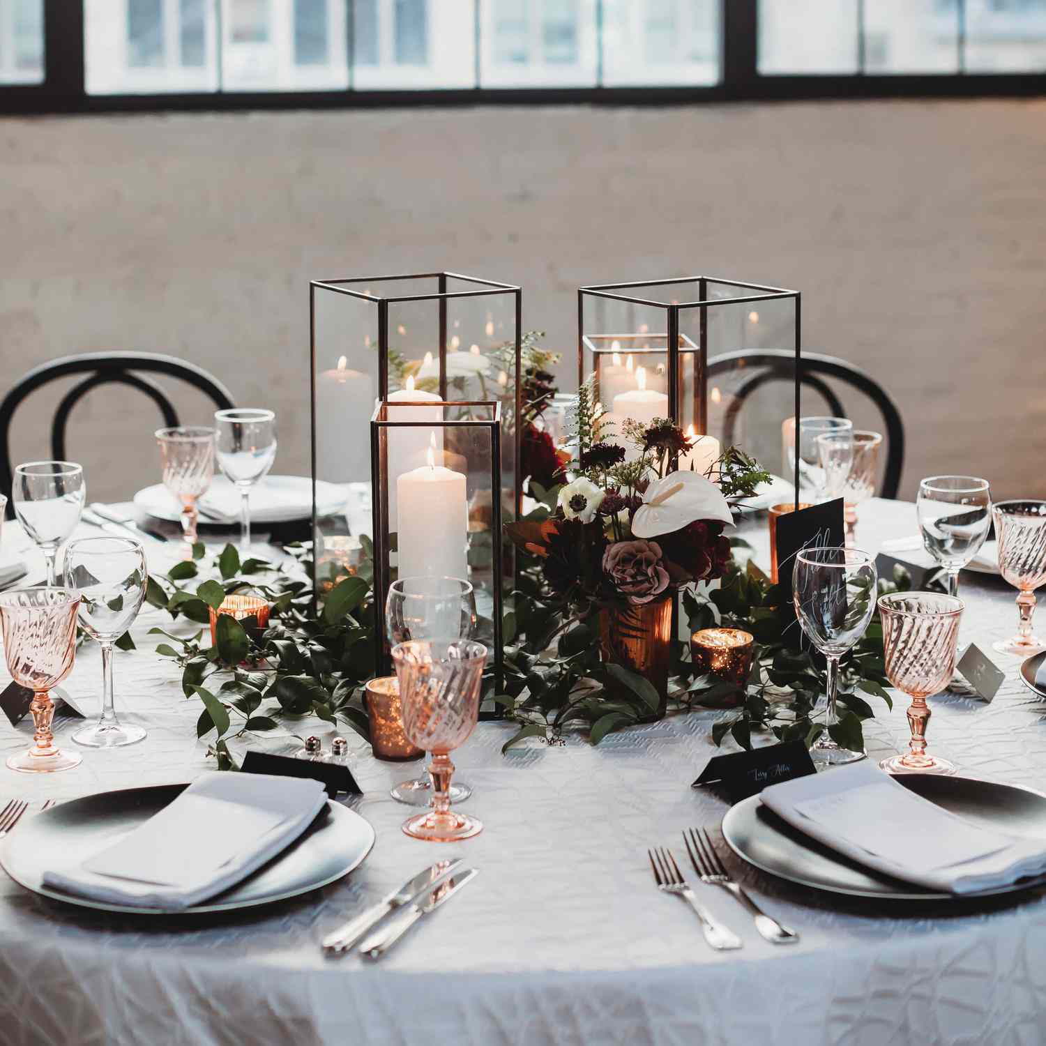 modern square black and glass lantern centerpieces atop greenery, white, and red flowers, at a table set with metallic dining ware.