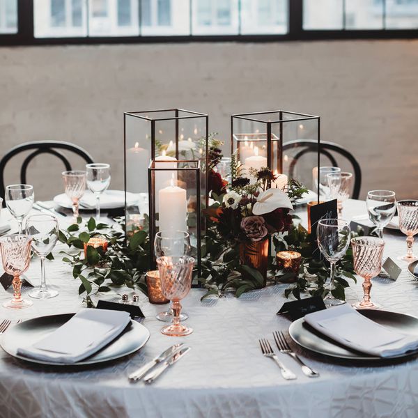 modern square black and glass lantern centerpieces atop greenery, white, and red flowers, at a table set with metallic dining ware.