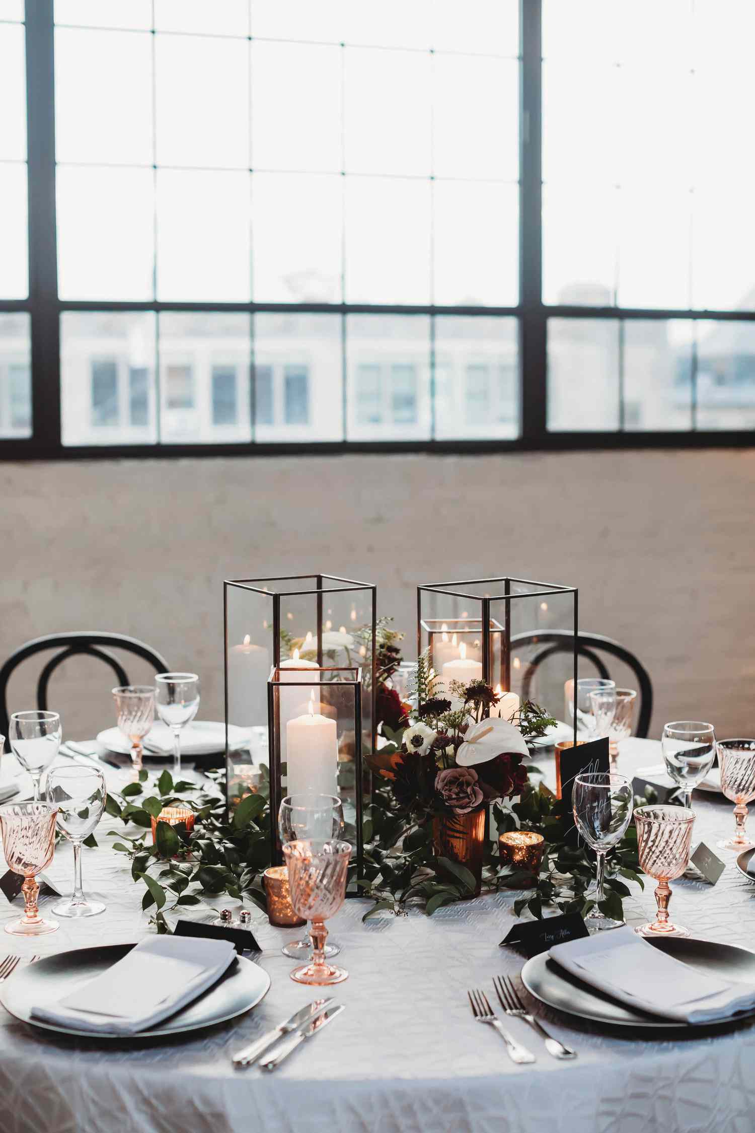 modern square black and glass lantern centerpieces atop greenery, white, and red flowers, at a table set with metallic dining ware.