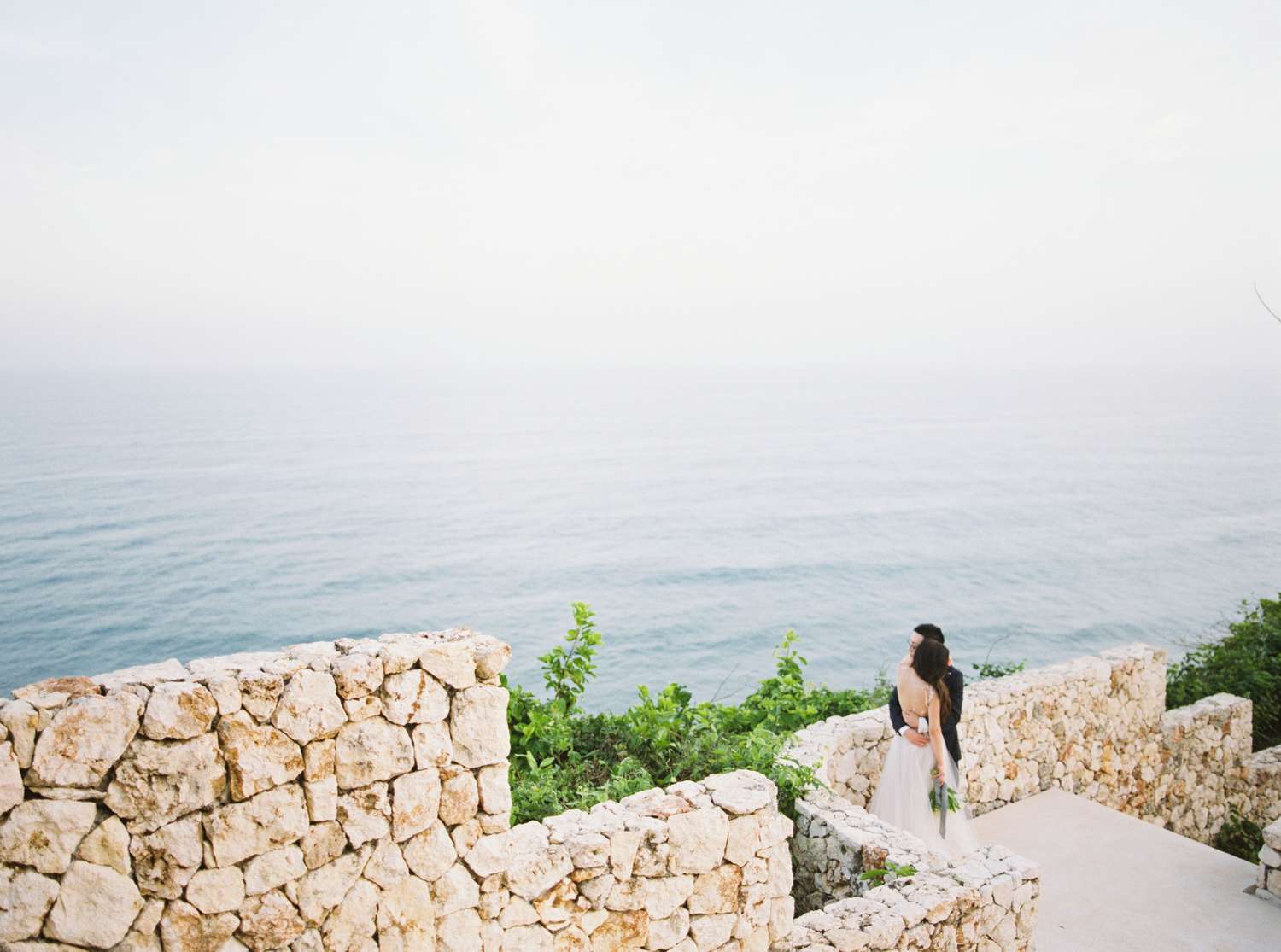 A bride and groom embrace on a cliff in Bali