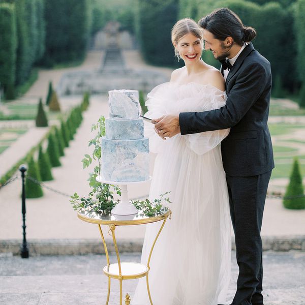 bride in off the shoulder tulle gown and groom in black tuxedo cutting into blue and white tiered wedding cake at garden estate