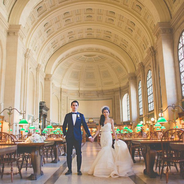 Bride and groom holding hands in the Boston Public Library, surrounded by wooden tables and green lamps during their wedding.