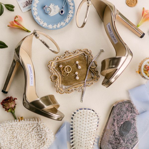 flat lay of wedding bridal items: gold heel sandals, jewelry, a hair brush, and flower petals
