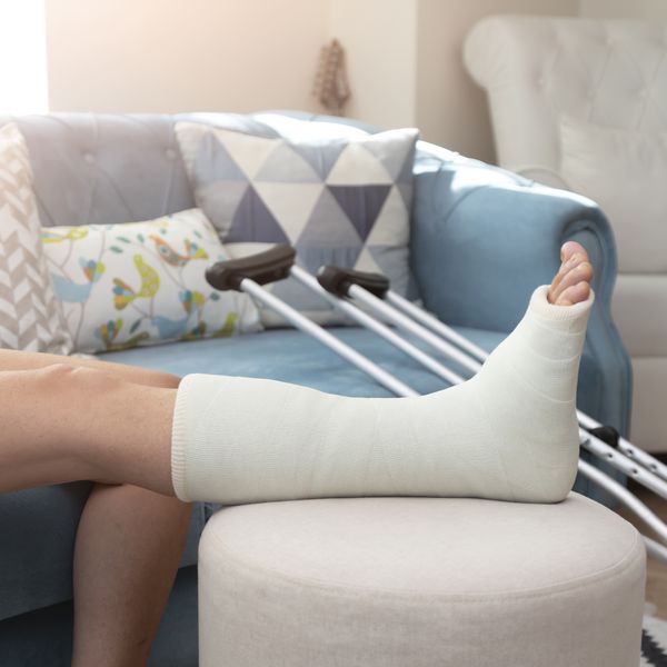 A personâs leg in a cast propped up on an ottoman with crutches lying on a blue couch in the background