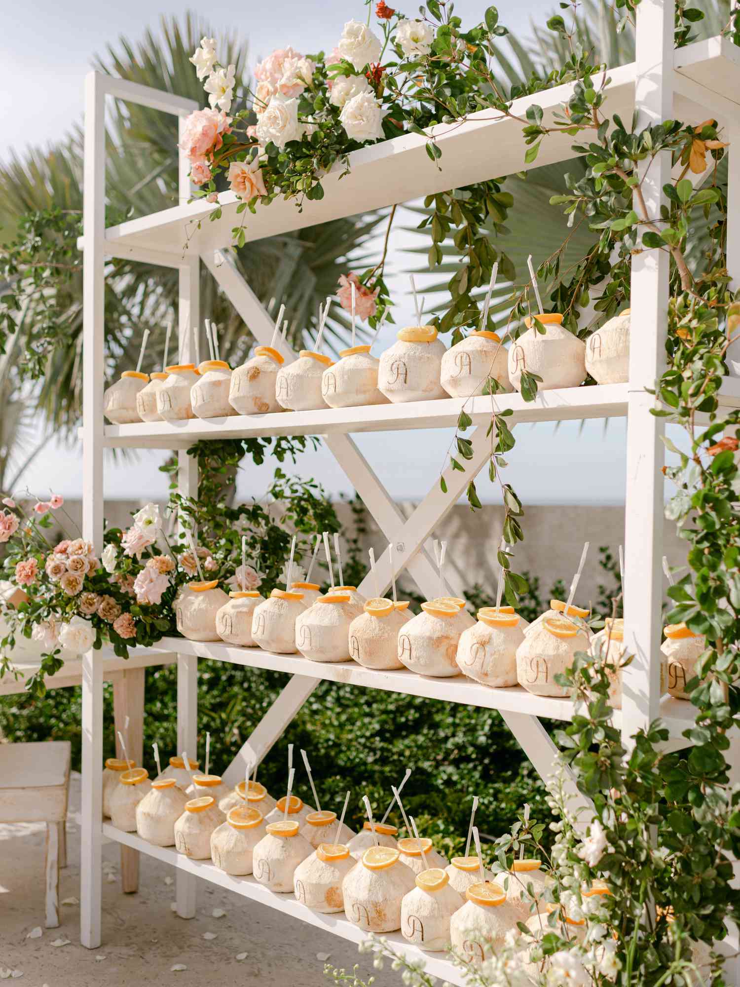 White shelving with monogrammed coconut escort cards with orange slices and straws framed with pink and white florals
