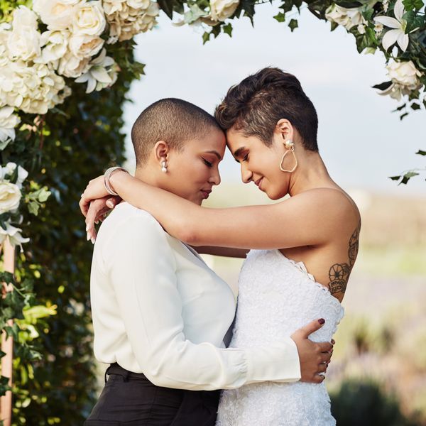 Two women stand under an arch of white roses at their wedding.