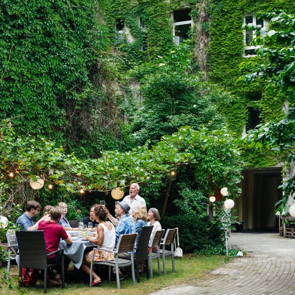 Family dinner outside of ivy-covered building in a courtyard with family members seated around table while father gives toast