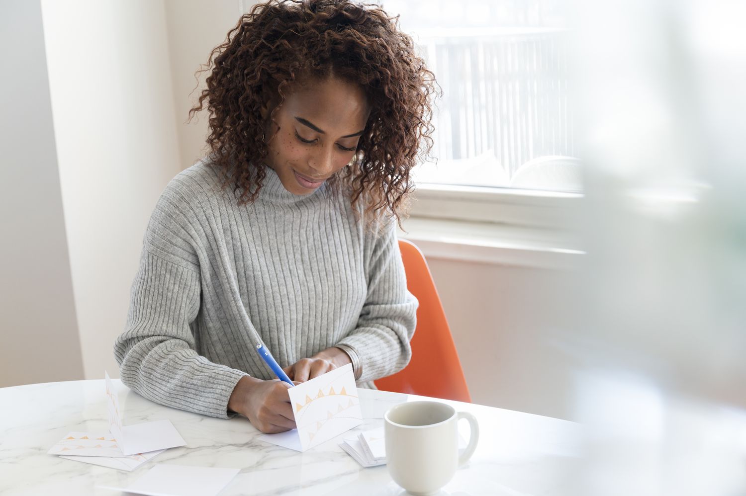 Young black woman seated at a table writing a wedding thank-you card while drinking coffee.