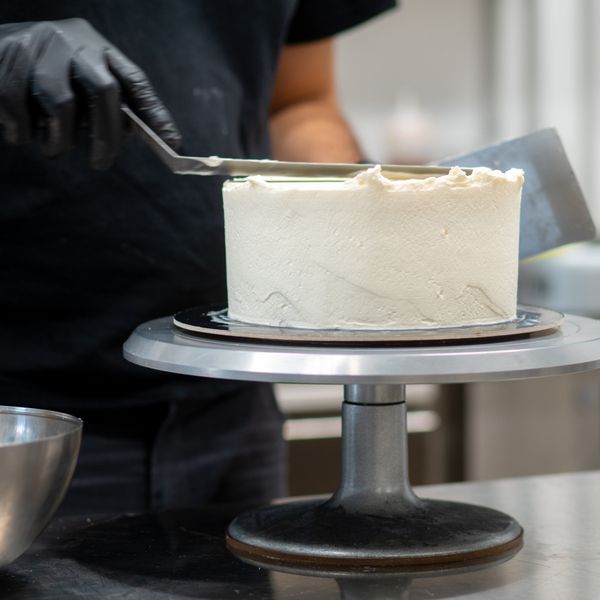 Closeup of Man in Black Gloves and Apron Spreading Buttercream on One Tier of Cake