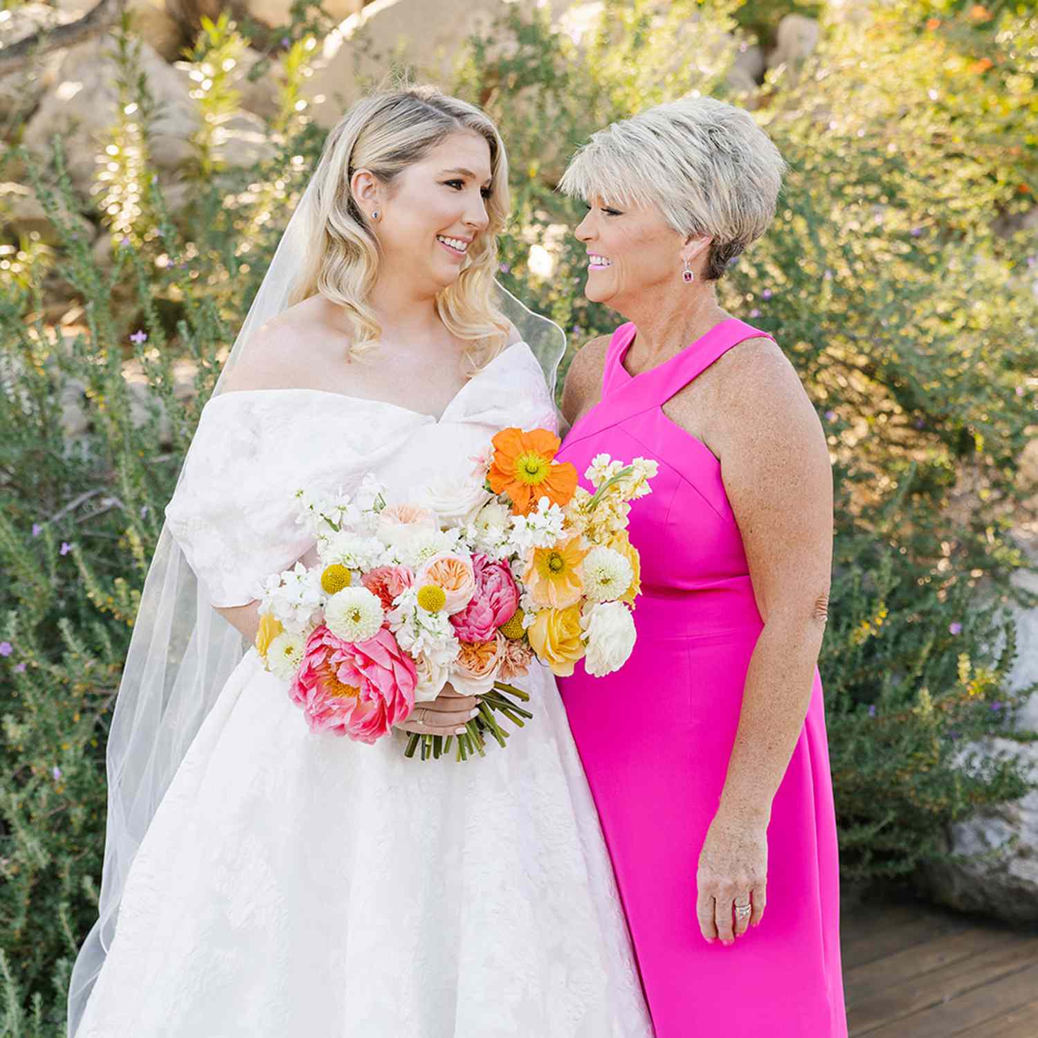 Bride in Off-the-Shoulder Wedding Dress Smiling at Mother of the Bride in Hot Pink Halter Neck Gown