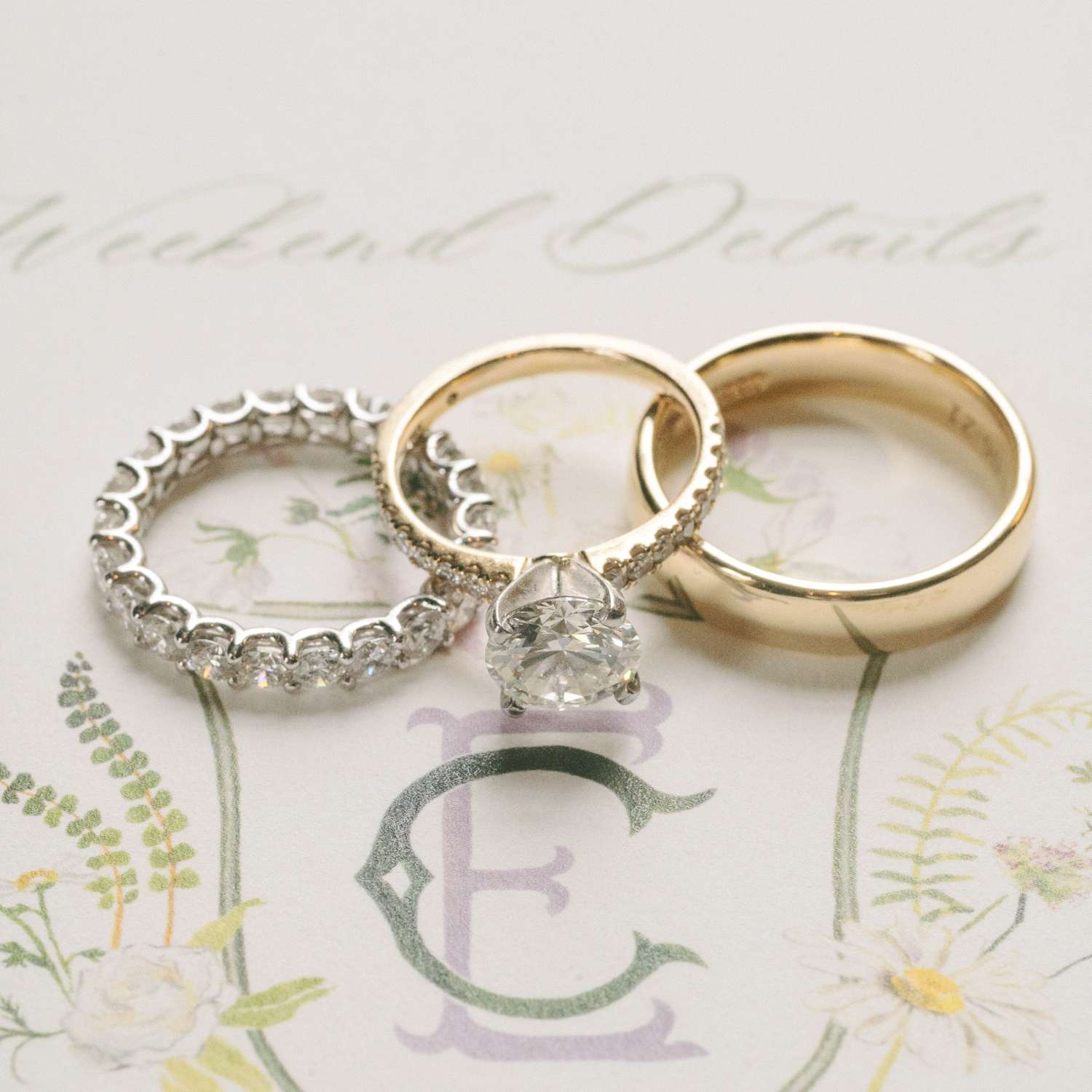 close up of a trio of rings: silver diamond eternity band; solitaire gold diamond engagement; gold wedding band