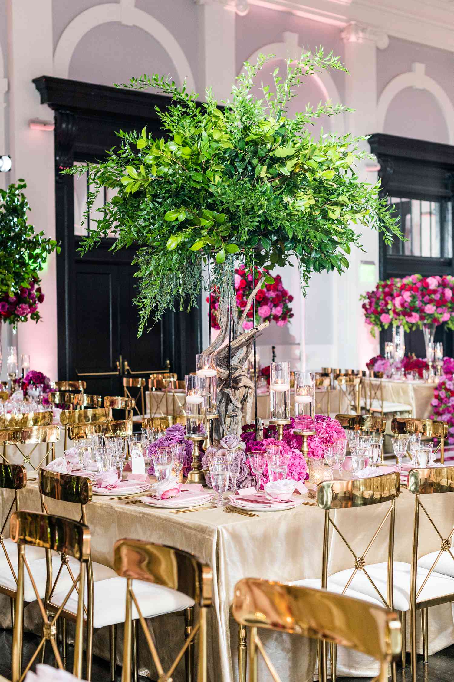 Tall tree centerpiece amid pink flora and gold chairs at reception hall.
