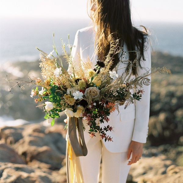 bride wearing white blazer holding colorful dried flower bouquet at an outdoor wedding.