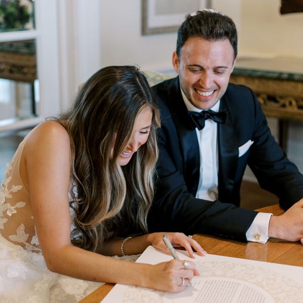 Bride in Floral Wedding Dress and Long, Wavy Hair Signing Ketubah While Groom in Tuxedo Smiles Next to Her