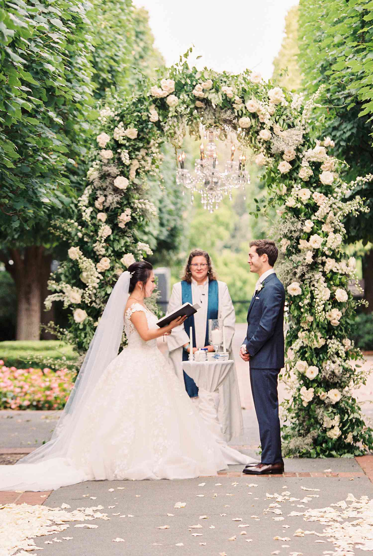 couple exchanging wedding vows under a flower arch with a hanging chandelier