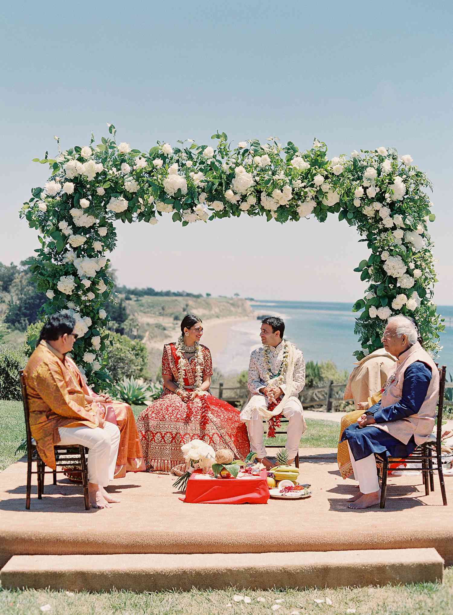 bride and groom sitting under a white floral mandap with their parents