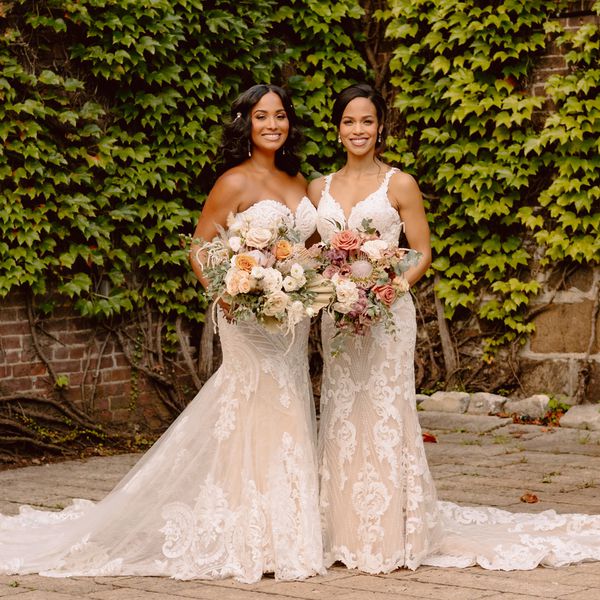 Two brides smiling in lace wedding dresses, one strapless mermaid and one trumpet with a V-neck