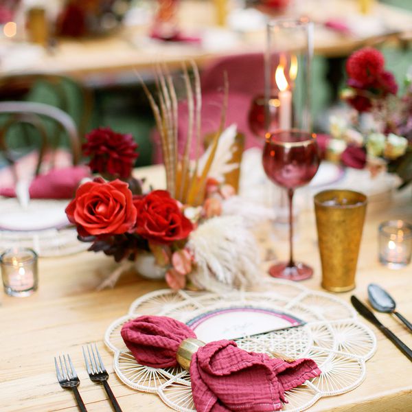 Place Setting With Rattan Charger, Burgundy Napkin, Black Flatware, Burgundy and Gold Glasses, and Red Rose Centerpieces on Wood Table Outside