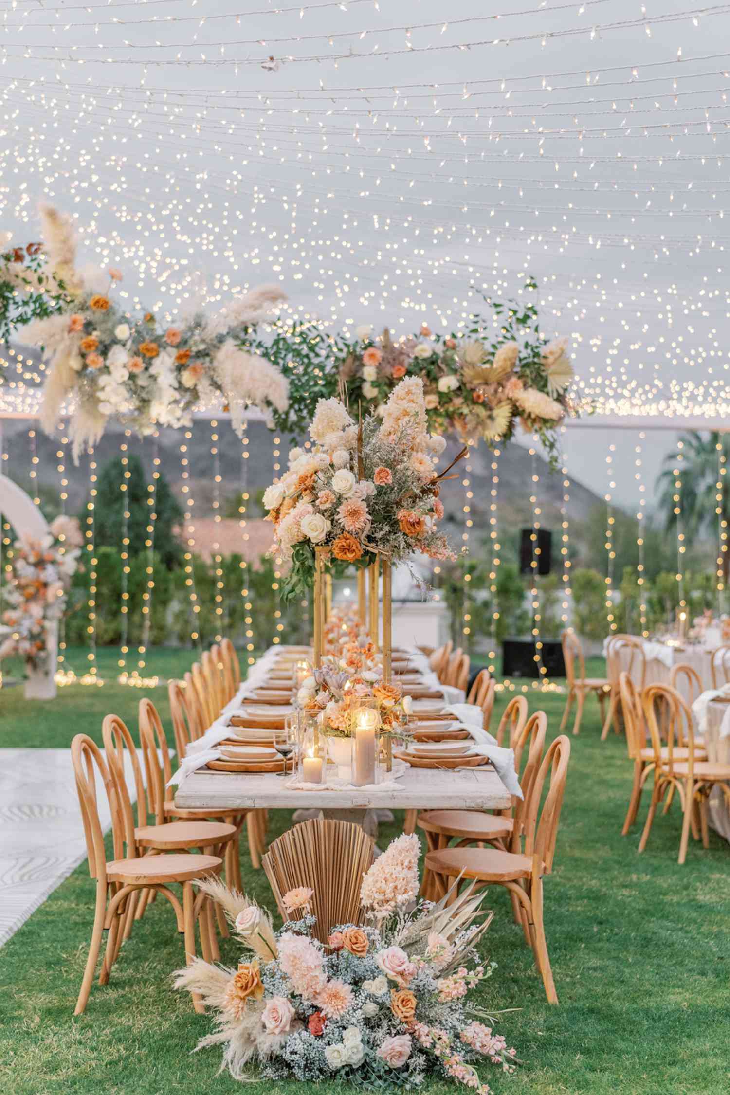 A tented wedding reception layout with long tables, wooden chairs, string lights, and floral centerpieces.