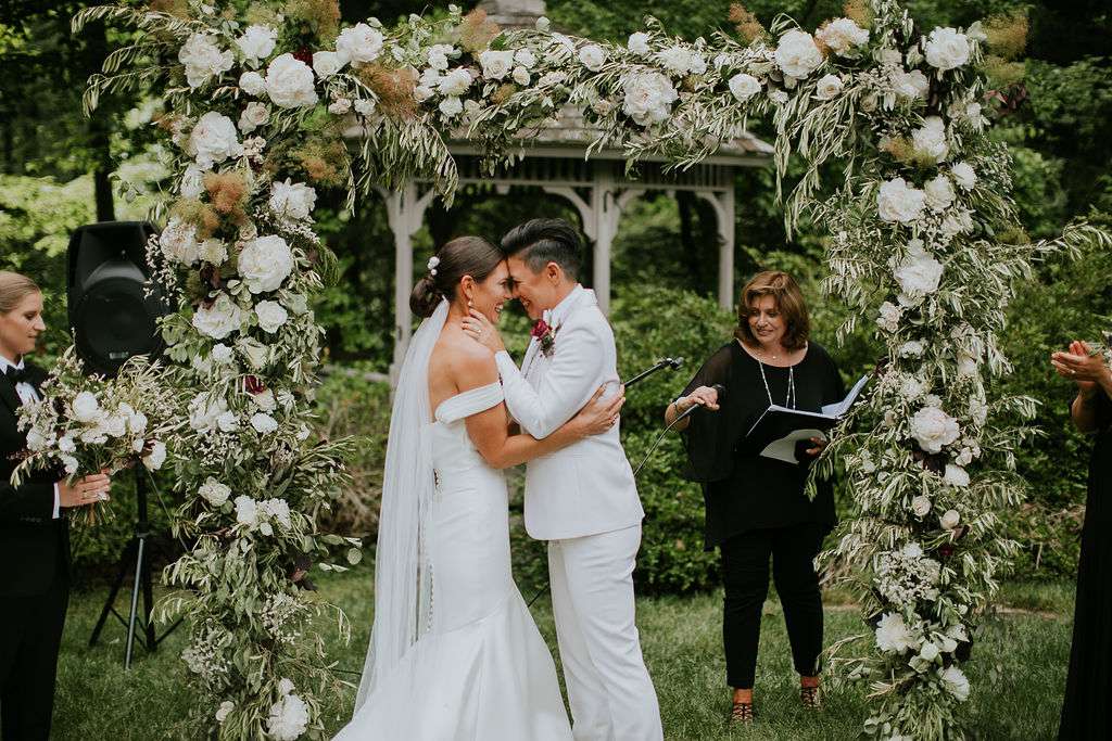 Two brides embracing in front of floral altar after wedding ceremony