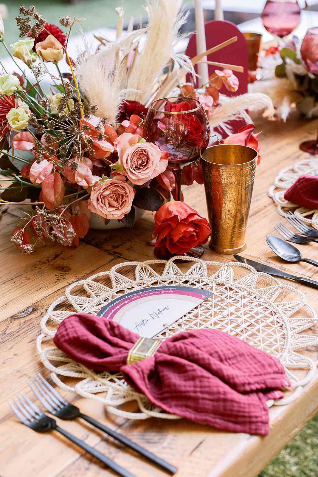 Place settings with rattan chargers, burgundy napkins, black flatware, and pampas grass centerpieces