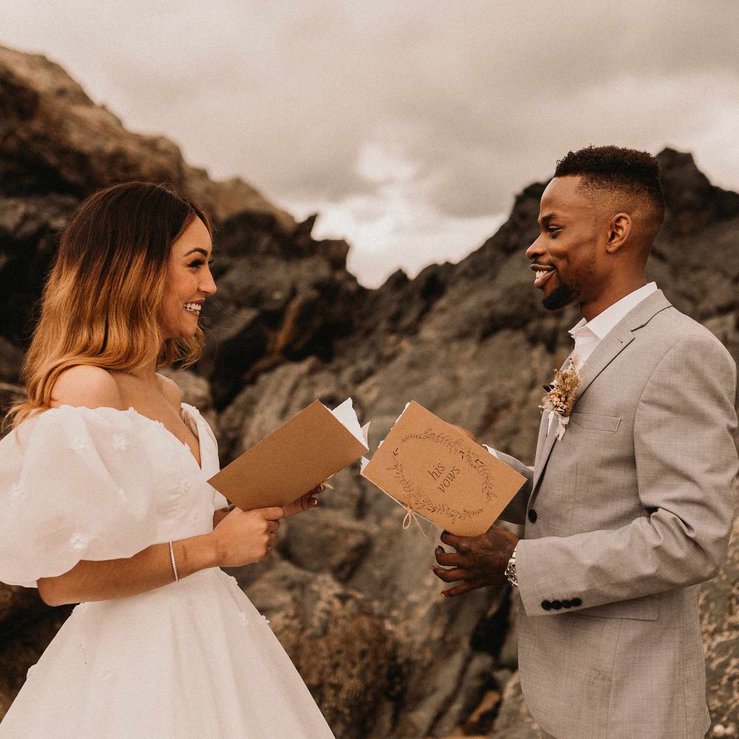 Smiling Bride and Groom Holding Vow Booklets With Mountain Backdrop Behind Them