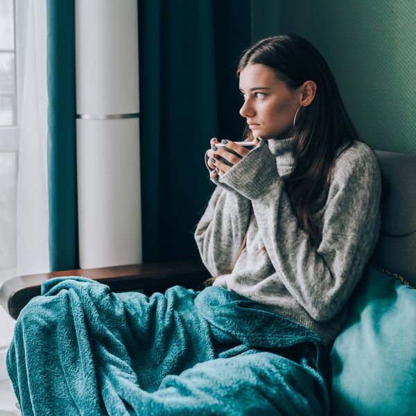Pensive, frost brunette woman in sweater, wrapped in green blue duvet sit on sofa and drink hot tea