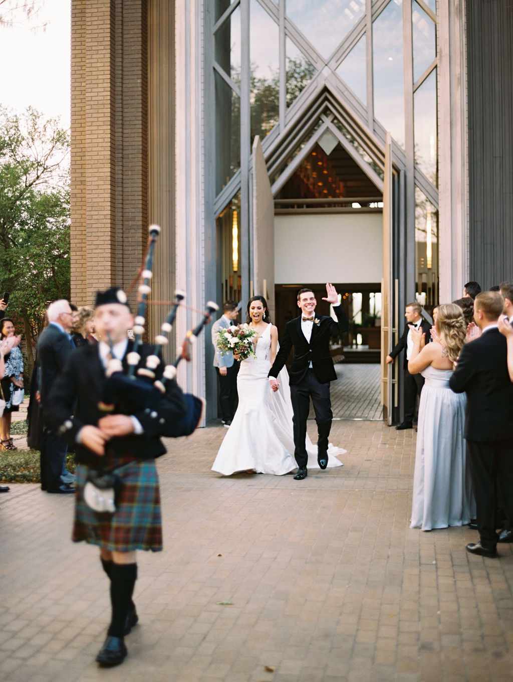 Bride and Groom Waving as They Leave the Church as Bagpipe Player Leads Them