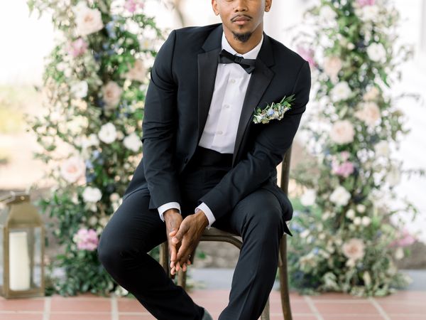 groom sitting on a chair while posing for a wedding portrait in front of a floral background, wearing a black tuxedo and a bowtie