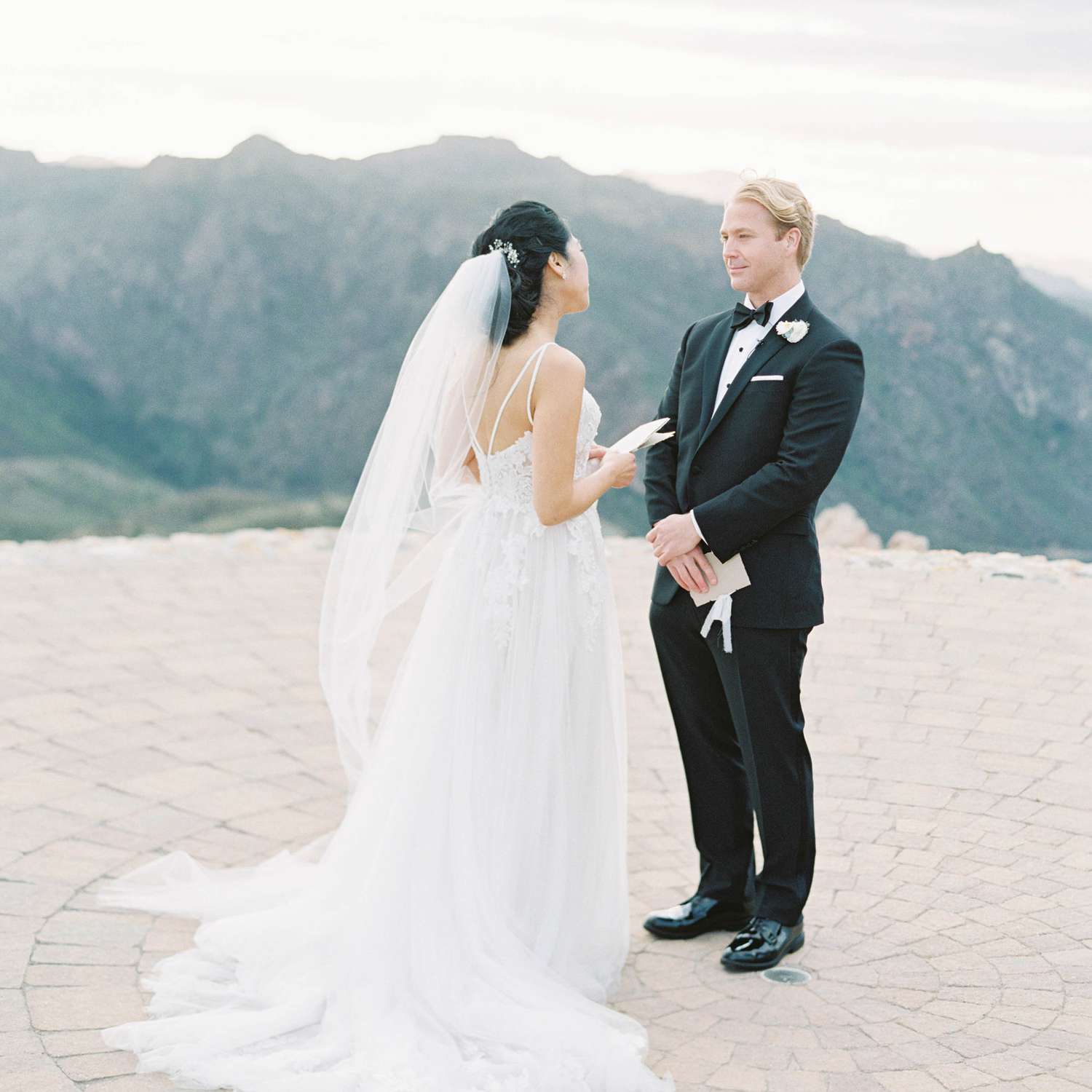 Back of Brunette Bride on Cliff Overlooking Mountains While Holding Booklet and Blonde Groom Gazing into Her Eyes
