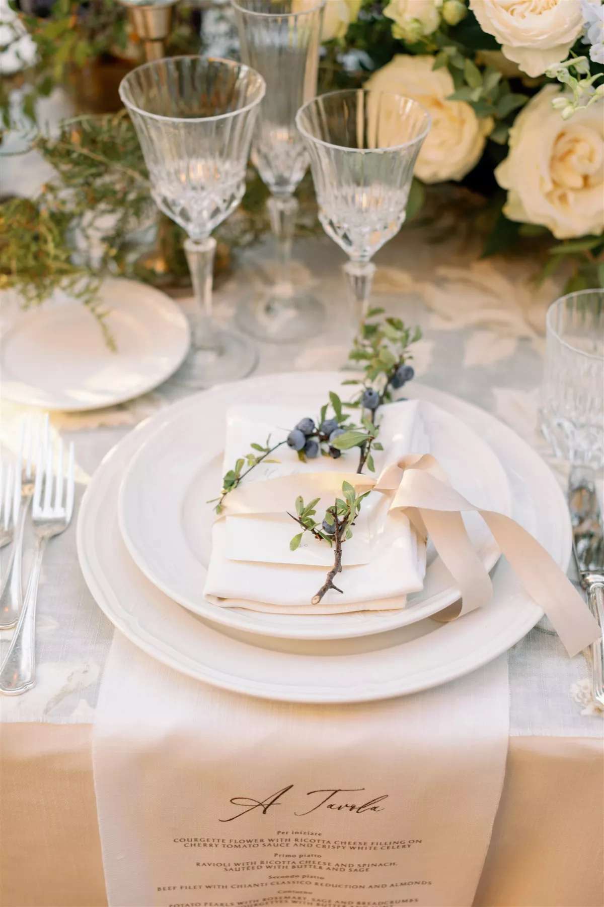 Wedding place setting with custom napkin menu, napkin tied with pink ribbon, greenery, and floral arrangements