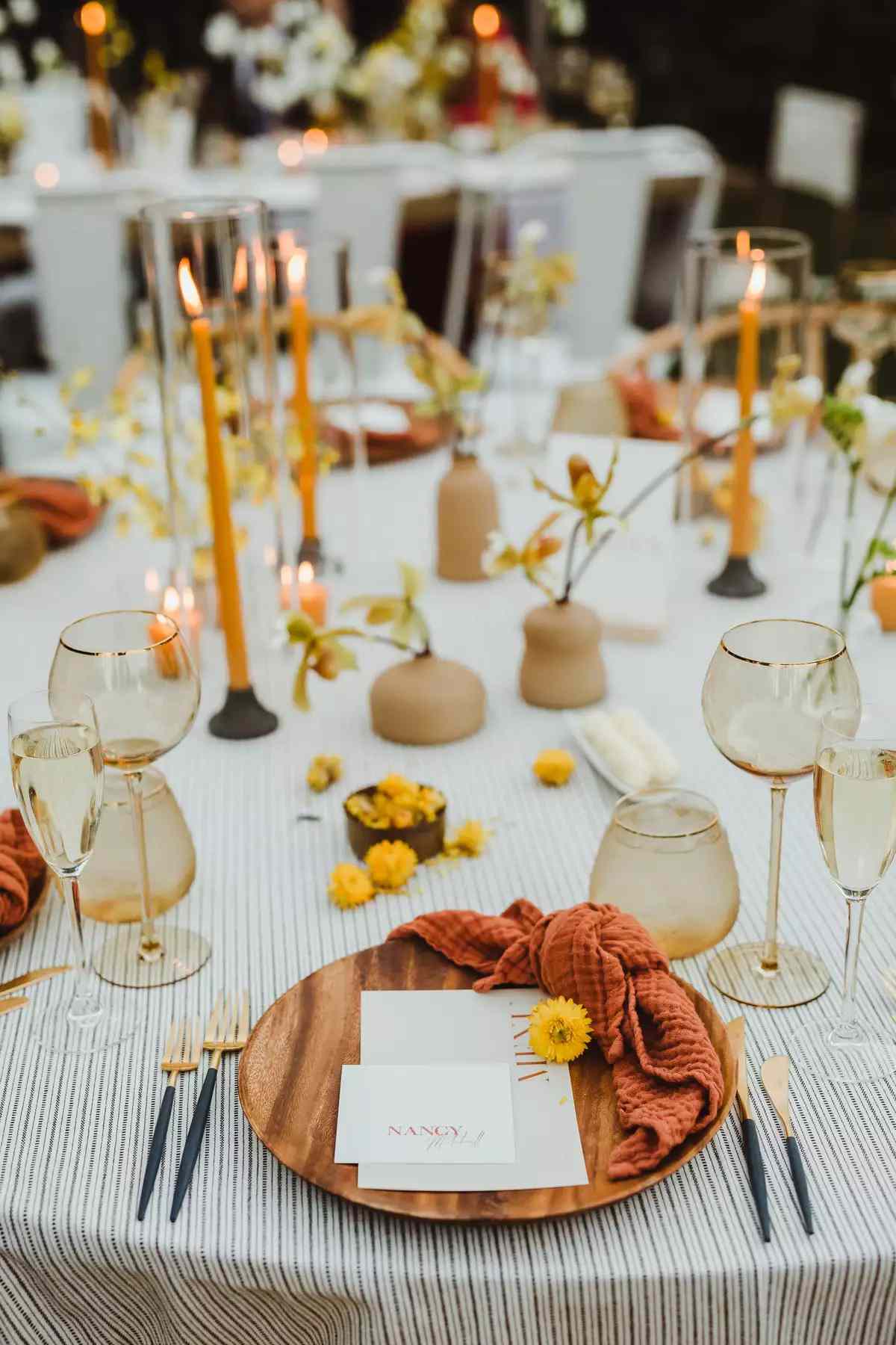 Wedding tablescape with orange candles, bud vases, yellow florals, wood plates, and orange napkins
