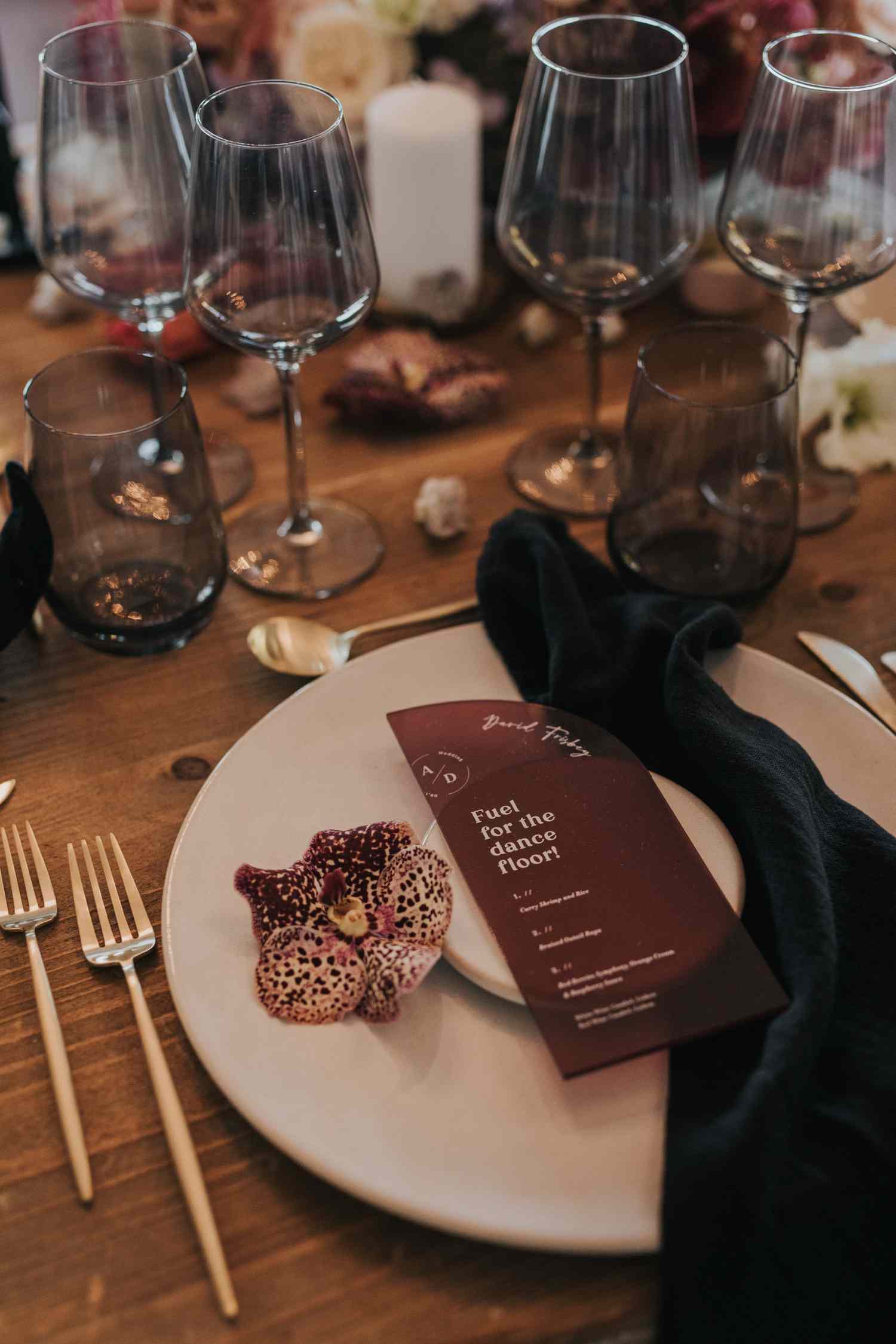 Wood tablescape with tinted glassware, white plate, dark napkin, burgundy menu, and an orchid