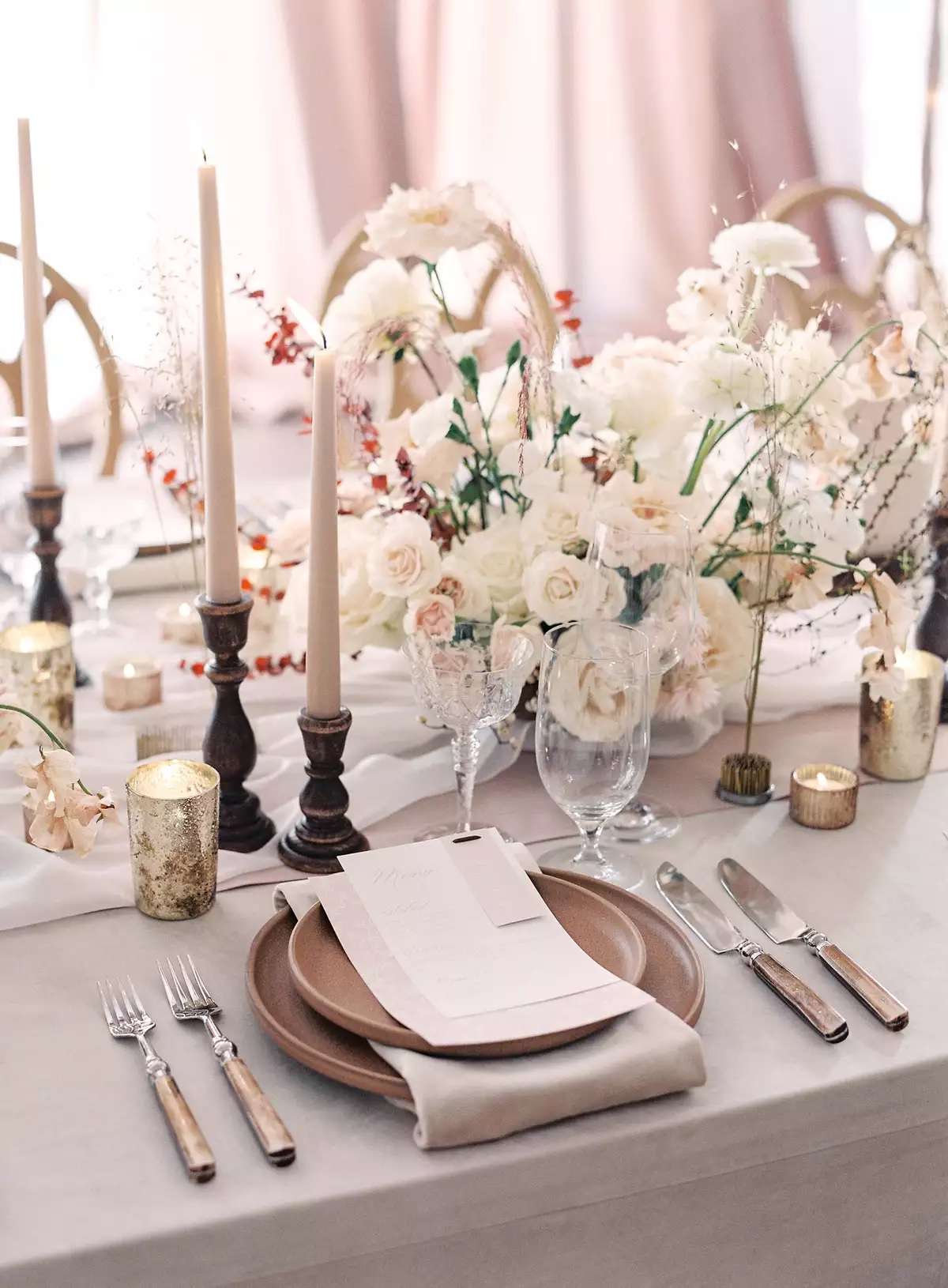 Tablescape with earth-tone plates, a pink and white runner, taper candles, white and red floral arrangements, and wood accented flatware