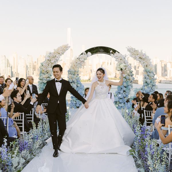 Couple walking up their aisle against backdrop of New York City skyline and floral arch and aisle florals with purple delphinium