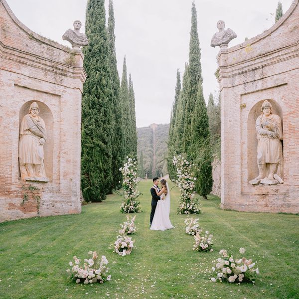 Bride in Wedding Dress and veil Posing With Groom in Black Tuxedo at Outdoor Altar Next to White Flower Decor and Classic Brick Architecture