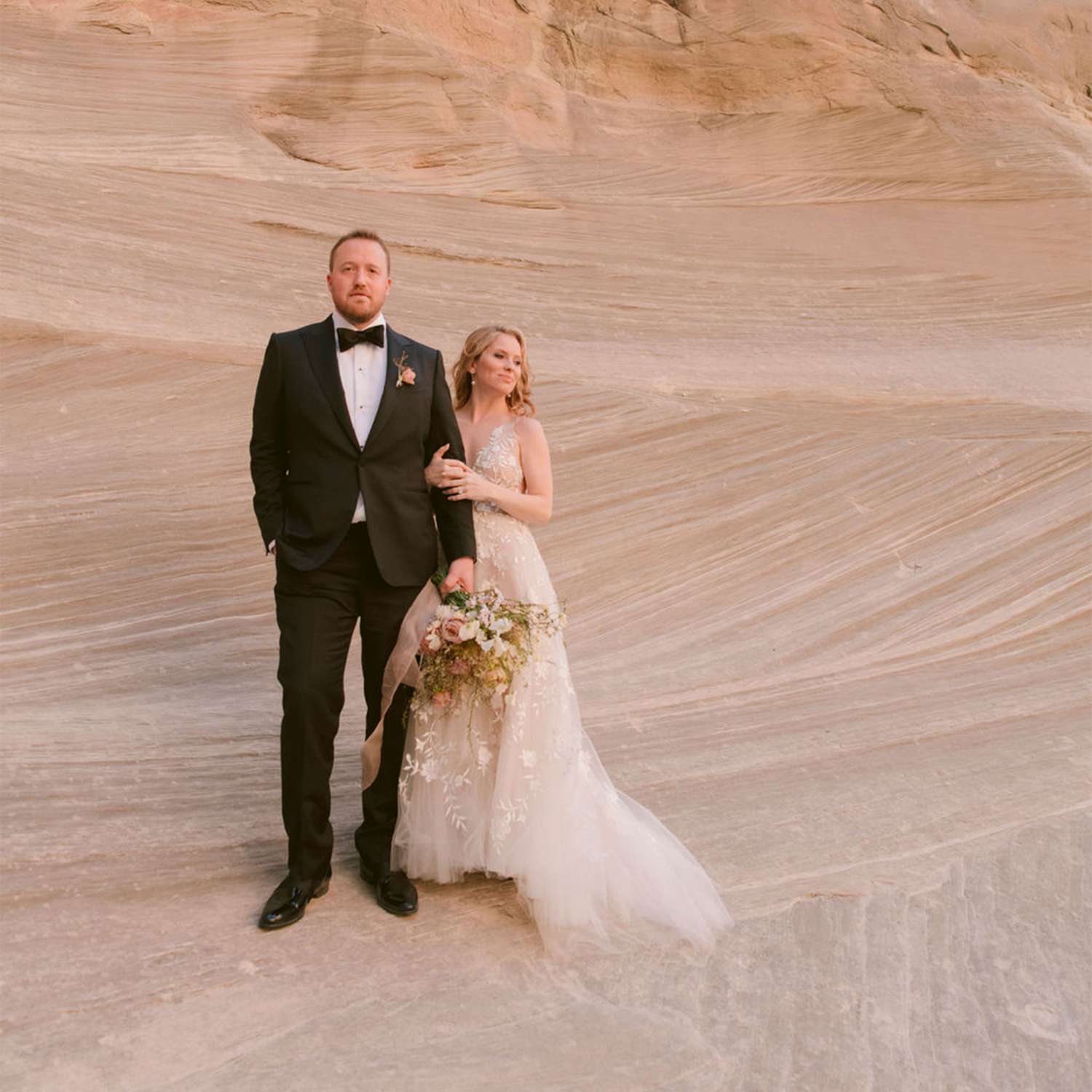 Bride in Pastel Wedding Dress Posing With Groom in Black Tuxedo in Front of Rock Formation