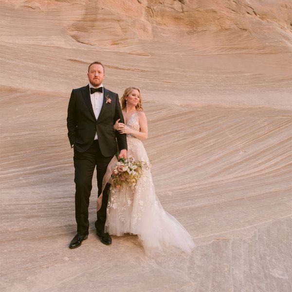 Bride in Pastel Wedding Dress Posing With Groom in Black Tuxedo in Front of Rock Formation