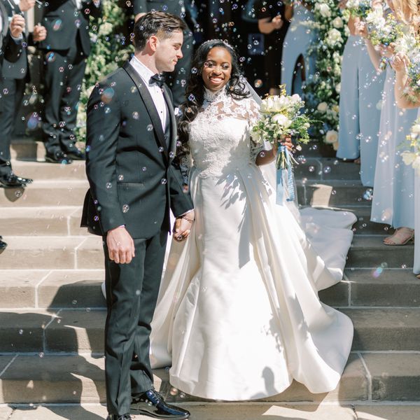 Bride in Long Sleeve Lace Wedding Dress Posing With Groom in Black Tuxedo In Front of Church While Guests Blow Bubbles