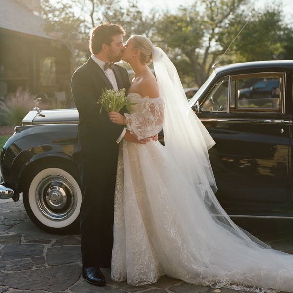 Bride in Strapless Lace Wedding Dress and Veil Kissing Groom in Black Tuxedo in Front of Black Classic Car