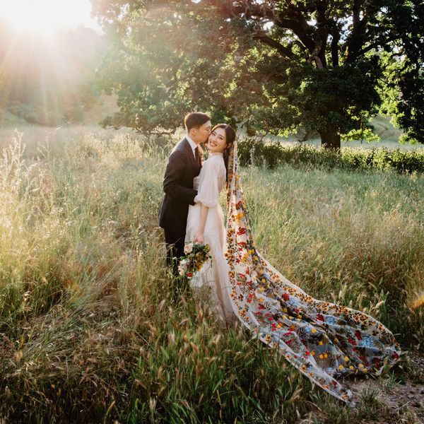 Groom in Black Suit Kissing Bride in White Dress and Colorful Floral Veil in Grassy Field