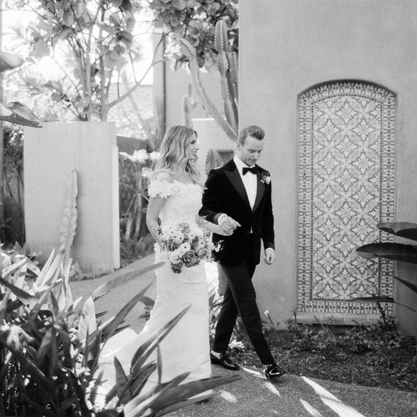 Black and White Image of Bride in Off the Shoulder Wedding Dress Walking With Groom in Tuxedo in Garden