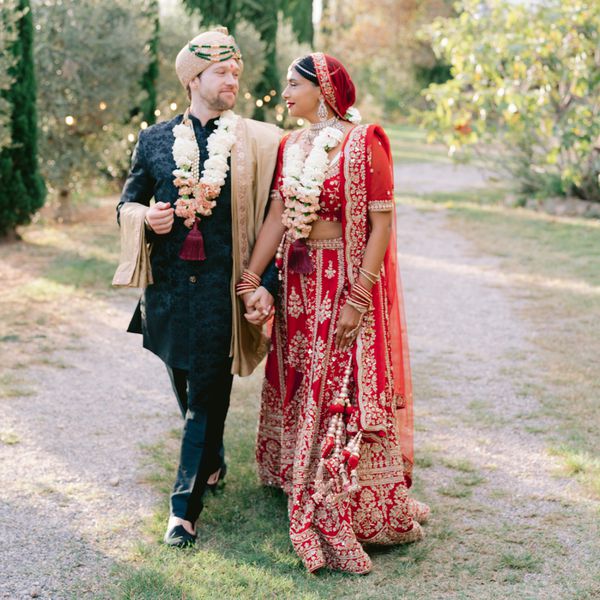 Bride in traditional Indian red dress with white flowers around neck holding hands with groom in black kurta and beige accessories