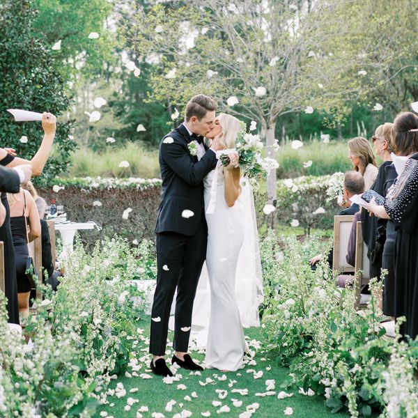Bride in Wedding Dress Kissing Groom in Black Tuxedo in Aisle With White Flower Petals Being Thrown