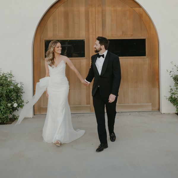 Erica Markle In Strapless Wedding Dress With Bow & Matt Romano in Black Tuxedo in Front of Arched Wooden Door