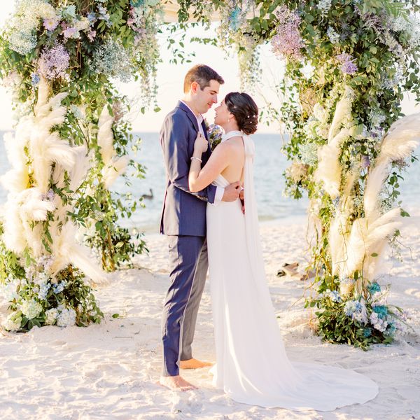 Bride in Halter Top Wedding Dress With Groom in Blue Suit In Front of Floral Altar in Front of Ocean