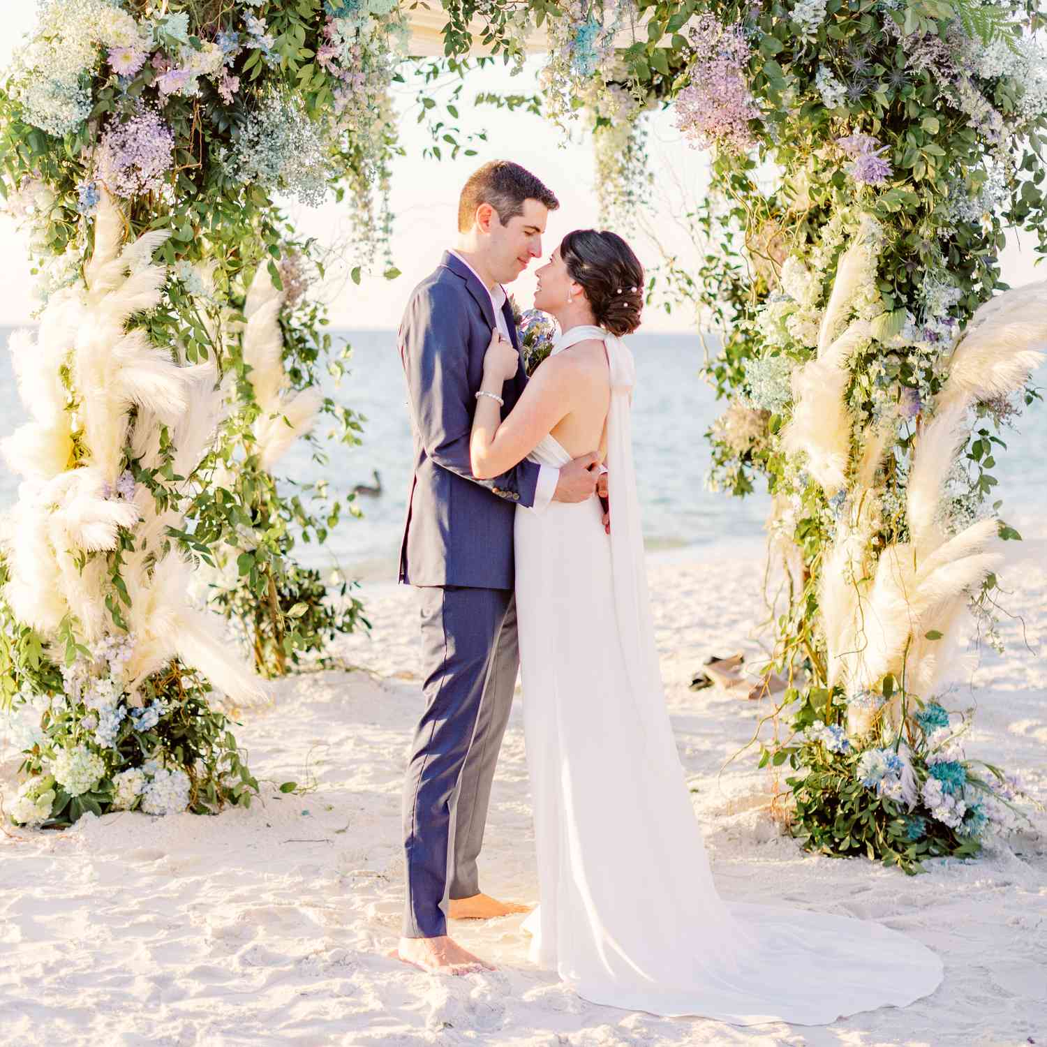 Bride in Halter Top Wedding Dress With Groom in Blue Suit In Front of Floral Altar in Front of Ocean