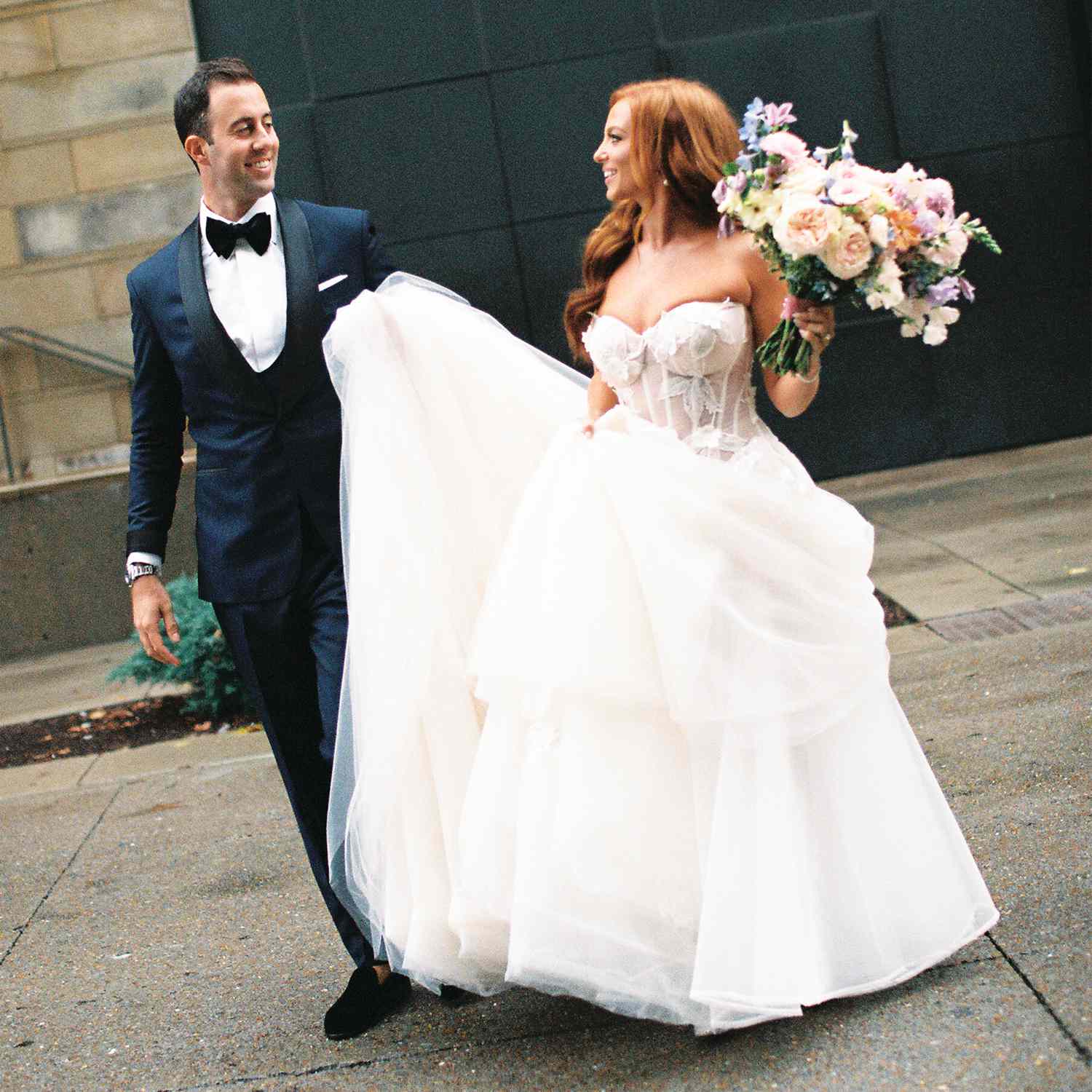 Bride in Strapless Wedding Dress Holding Bouquet Posing With Groom in Navy Tuxedo