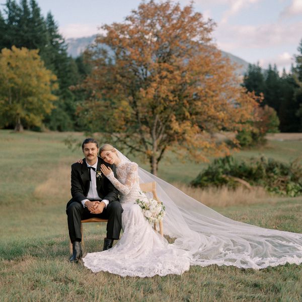 Bride in Lace Wedding Dress With Long Veil Seated With Groom in Black Tuxedo in Grass Field