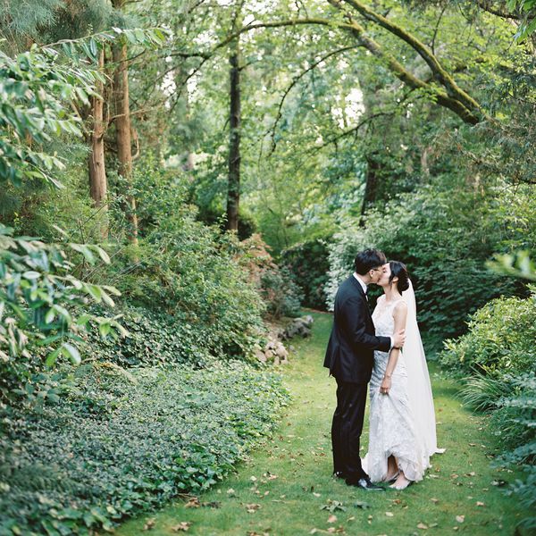 Bride in Wedding Dress With Veil Kissing Groom in Black Tuxedo in Wooded Area
